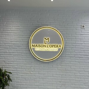 May include: A circular illuminated sign with the words "MAISON L'OPERA RESTAURANT & P&Acirc;TISSERIE" in gold. The sign has a gold border and a grey background, mounted on a white brick wall.