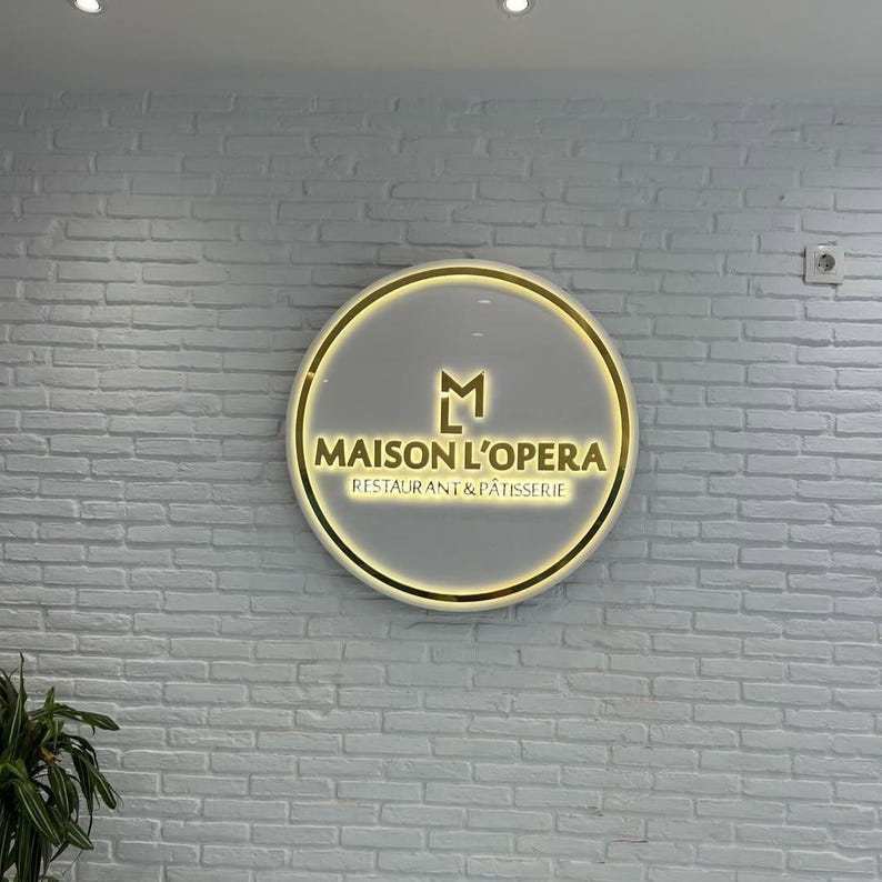 May include: A circular illuminated sign with the words "MAISON L'OPERA RESTAURANT & P&Acirc;TISSERIE" in gold. The sign has a gold border and a grey background, mounted on a white brick wall.