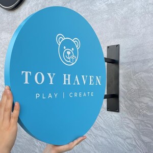 May include: A round, light blue sign with the text "TOY HAVEN" and the words "PLAY | CREATE" in white. A white bear logo is at the top. The sign is mounted on a black metal bracket.