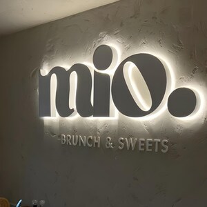 May include: A sign on a textured wall reads "mio." in a stylized font, with the words "BRUNCH & SWEETS" below. The sign is a neutral grey colour with a bright white glow around the letters.