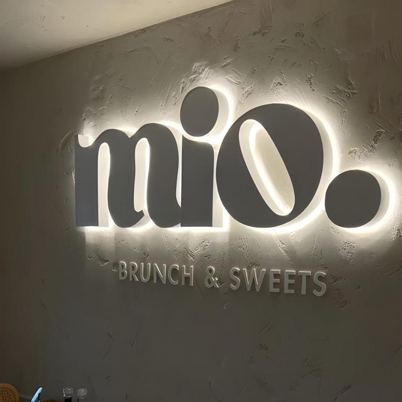 May include: A sign on a textured wall reads "mio." in a stylized font, with the words "BRUNCH & SWEETS" below. The sign is a neutral grey colour with a bright white glow around the letters.