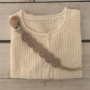 Könnte beinhalten: Cremefarbene Baby-Strickjacke mit Knopfleiste und zartem Lochmuster. Ein gehäkelter Schnullerclip in einem neutralen Farbton ist befestigt. Der hölzerne Schnullerhalter ist sichtbar. Die Strickjacke ist ordentlich auf einer hellen Holzoberfläche gefaltet.
