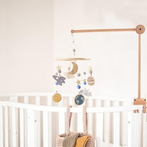Op de afbeelding: Een houten babymobiel met een hemels thema. Het heeft een halve maan, sterren, planeten, een raket en een pluche witte wolk. De mobiel hangt aan een houten arm die aan een wieg is bevestigd.
