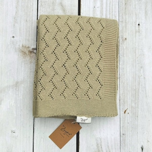 Peut inclure: Une couverture pour bébé en tricot beige avec un motif en losange. La couverture est pliée en deux et porte une étiquette brune avec le mot "Zaygoo" dessus.