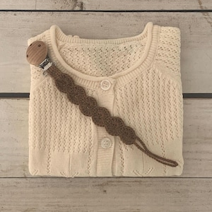 Könnte beinhalten: Cremefarbene Baby-Strickjacke mit Knopfleiste und dekorativem braunen Schnullerhalter. Die Strickjacke ist ordentlich gefaltet, und der Schnullerhalter besteht aus einem Holzclip und einem braunen gehäkelten Riemen.