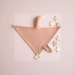 Può includere: Una coperta rosa per bambini a forma di oca, con un lungo collo e un becco bianco. La coperta ha un aspetto strutturato ed è circondata da decorazioni a forma di stella dai colori pastello. Lo sfondo è rosa chiaro.