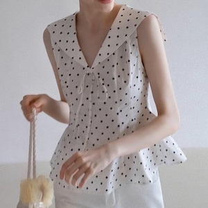Könnte beinhalten: Ein ärmelloses weißes Oberteil mit schwarzen Punkten und einem Bubikragen. Das Oberteil hat eine Bindedetail am Ausschnitt und einen Schößchensaum. Die Person trägt eine weiße Hose und eine passende Schleife mit Punkten im Haar. Sie hält eine kleine, gewebte Tasche.