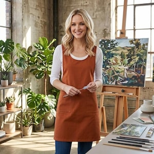 May include: A rust-colored apron with two front pockets is worn over a white long-sleeved shirt. The artist is holding a palette knife in a studio filled with plants, paintings, and art supplies.