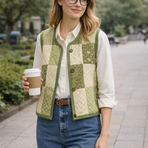 Könnte beinhalten: Gesteppte Weste mit Patchwork-Design in Grün- und Cremetönen, mit Knopfleiste und braunem Ledergürtel. Die Weste wird über einem weißen Hemd und einer blauen Jeans getragen. Die Person hält einen Kaffeebecher.