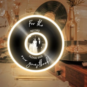 May include: A white acrylic sign with a black vinyl record design. The sign says "For the Record" and "Please Sign our Guestbook!" with a photo of a couple in the centre.