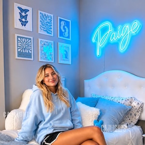 May include: A bright blue neon sign displaying the name Paige in a cursive script font, mounted on a gray wall above a white tufted headboard. The sign emits a vibrant glow, illuminating the bedroom space with a cool blue light.