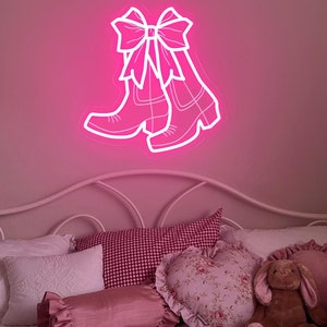 May include: A neon pink sign in the shape of cowboy boots with a bow, mounted on a pink wall. The sign is above a bed with white bedding and decorative pillows in shades of pink and red.
