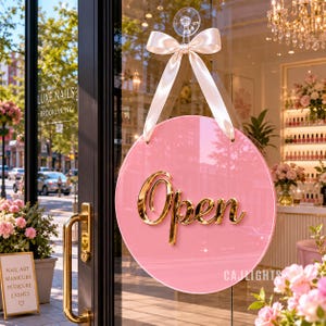 Op de afbeelding: Een rond roze bord met het woord Open in goudkleurige schrijfletters, opgehangen aan een crèmekleurig satijnen lint met strik aan een doorzichtige zuignap op een glazen deur. Het bord heeft een glanzende afwerking en reflecteert de omgeving.