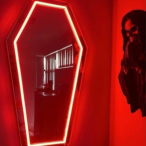 May include: A coffin-shaped mirror with a bright red neon border. The mirror is mounted on a red wall, and a decorative skull figure is to the right. The room is dimly lit, creating a spooky atmosphere.