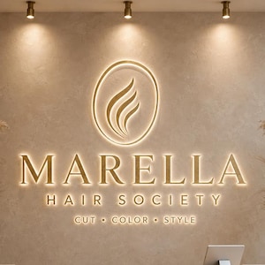 May include: A beige reception desk with a wall sign featuring the gold backlit text MARELLA HAIR SOCIETY, CUT COLOR STYLE, and a flame-like logo inside an oval. Two large beige vases with dried pampas grass flank the desk, and a tablet sits on top.