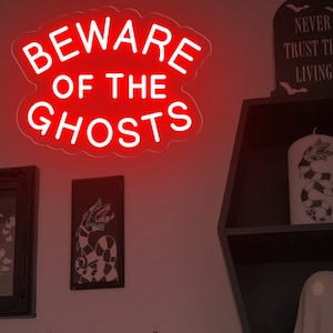 Op de afbeelding: Een rood neon-bord met de woorden "Beware of the Ghosts" op een witte muur. Het bord is omringd door Halloween-decoraties, waaronder een geest, een grafsteen en een ingelijst afdruk.