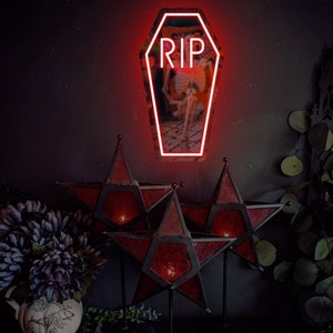 May include: Red neon sign shaped like a coffin with the word "RIP" inside. The sign is hanging on a dark wall with three red star-shaped lanterns in the foreground.