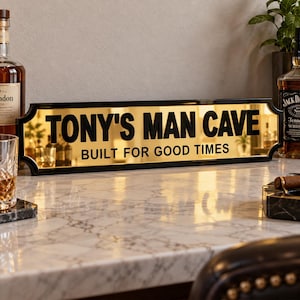 以下が含まれることがあります： 黒いフレームが付いた長方形のミラーサイン。太い黒文字で「TONY'S MAN CAVE」および「BUILT FOR GOOD TIMES」と記されています。反射面はゴールド調の仕上げで、ホームバーエリアの装飾品として機能します。