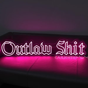May include: White neon sign with the words "Outlaw Shit" in a gothic font. The sign is lit up in a bright pink color and is sitting on a pink couch.