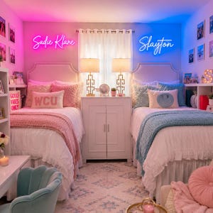 May include: Two twin beds with white bedding, pink and blue textured throws, and decorative pillows. Above the beds are custom neon signs reading Sadie Klane in pink and Slayton in blue. A white cabinet with two lamps sits between the beds.