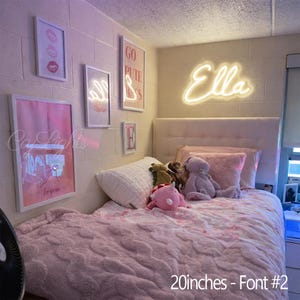 May include: A custom neon sign displaying the name Ella in a cursive script font, mounted on a white cinder block wall above a bed. The sign measures 20 inches in width. The bedroom features pink-toned wall art and a textured light pink blanket.