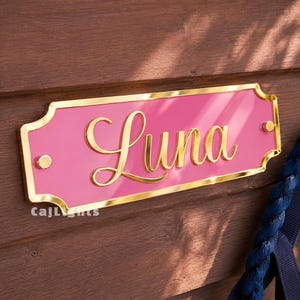 May include: A rectangular pink sign with a gold-colored border and gold-colored script lettering that reads Luna. The sign features rounded corners with decorative indentations and two gold-colored circular mounting points on the left and right sides.