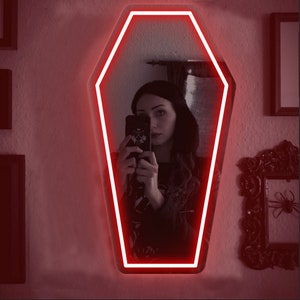 May include: Coffin-shaped mirror with a bright red neon outline. The mirror reflects a person holding a phone. The wall is painted a light color, and there are framed pictures on the wall.