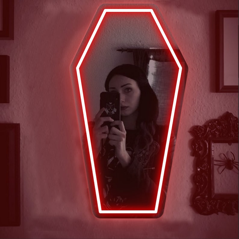 May include: Coffin-shaped mirror with a bright red neon outline. The mirror reflects a person holding a phone. The wall is painted a light color, and there are framed pictures on the wall.