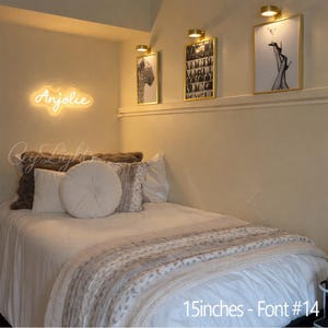 May include: A warm yellow neon sign displaying the name Anjolie in a cursive script font, mounted on a wall above a bed. The sign measures 15 inches in width and uses font style number 14, creating a soft glow in the bedroom space.