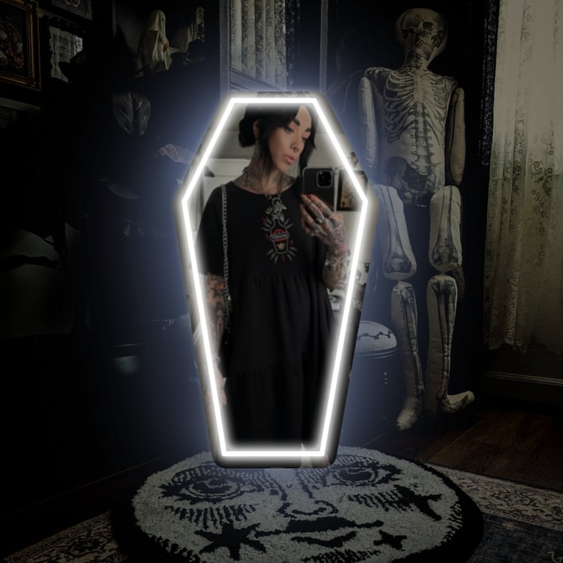 May include: A coffin-shaped mirror with a bright white neon outline. The mirror reflects a person in a black dress. A decorative rug with a moon face design is on the floor. A skeleton decoration stands in the background.