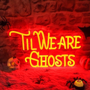 May include: Yellow neon sign with the text "Til We Are Ghosts" on a red brick wall background. The sign is lit up and there are Halloween decorations around it.