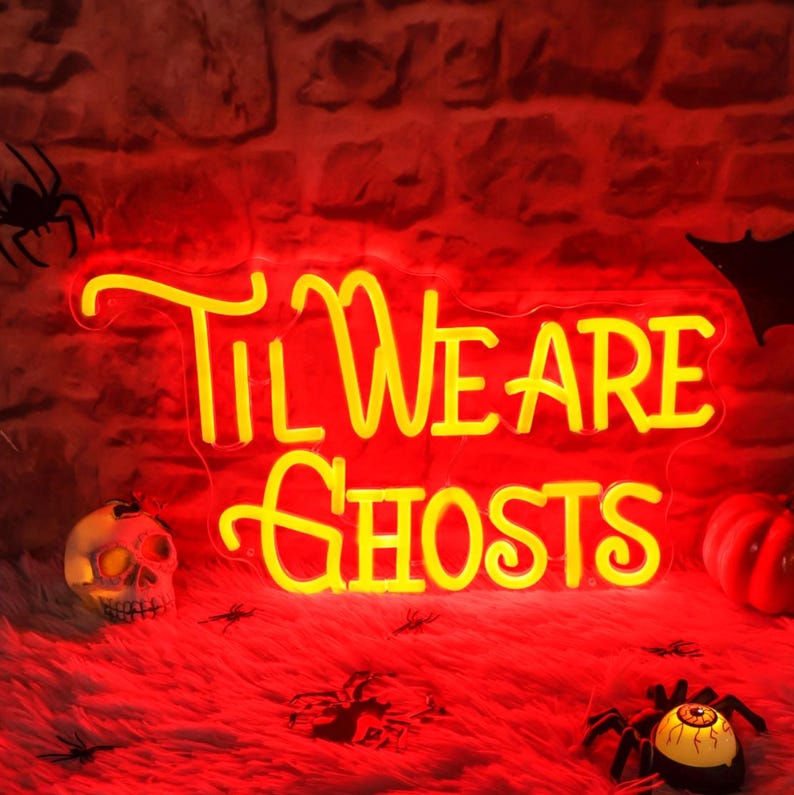 May include: Yellow neon sign with the text "Til We Are Ghosts" on a red brick wall background. The sign is lit up and there are Halloween decorations around it.