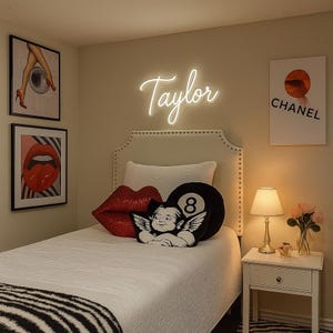 May include: A white neon sign displaying the name Taylor in a cursive script font mounted on a wall above a bed. The bed features a white quilt, a red sequined lip-shaped pillow, a black eight-ball pillow, and a black and white cherub pillow.