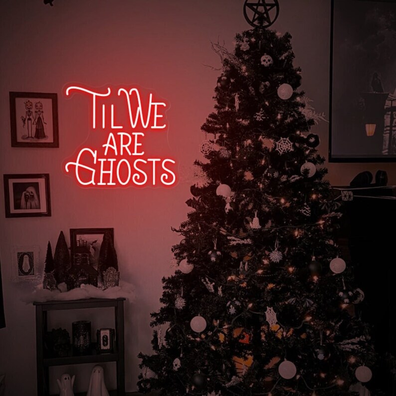 May include: A red neon sign with white lettering that reads "Til We Are Ghosts" hanging on a wall next to a black Christmas tree decorated with white ornaments and a star on top.