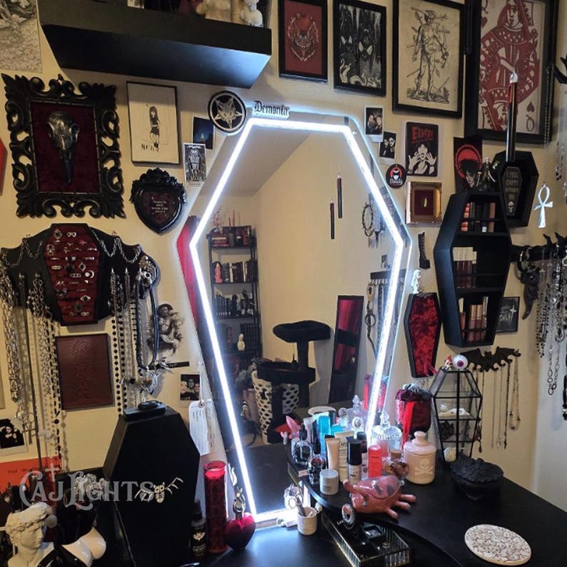 May include: A coffin-shaped mirror with a white neon outline is the focal point of a gothic-themed vanity. The black vanity is adorned with various gothic decor, including framed artwork, jewelry, and coffin-shaped shelves. The room is decorated in dark colors.