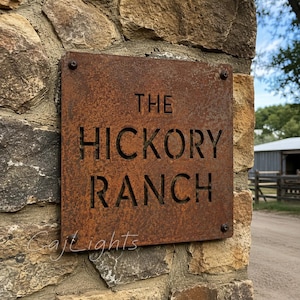 May include: A square, rusted metal sign with cut-out lettering that reads THE HICKORY RANCH. The sign is secured to a stone surface with four dark screws, one in each corner, and features a weathered, oxidized brown finish throughout the surface.