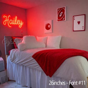 May include: A bedroom featuring a custom red neon sign displaying the name Hailey in cursive script. The bed is dressed in white linens with a red textured throw blanket, white pillows, and wall art depicting a red billiard ball and playing cards.