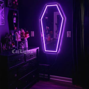 May include: A coffin-shaped mirror with a bright purple neon outline hangs on a dark purple wall. The mirror reflects a room with a dresser, shelves, and various decorative items. The text "CajLights" is visible on the dresser.