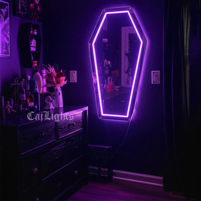 May include: A coffin-shaped mirror with a bright purple neon outline hangs on a dark purple wall. The mirror reflects a room with a dresser, shelves, and various decorative items. The text "CajLights" is visible on the dresser.