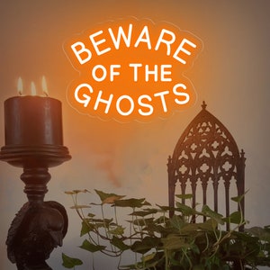 Op de afbeelding: Een neonbord met de tekst "Beware of the Ghosts" in een witte omtrek tegen een oranje achtergrond. Het bord hangt in een kamer met een donkere houten kandelaar en een zwarte metalen stoel met een gotische stijl.