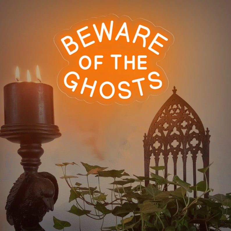 Op de afbeelding: Een neonbord met de tekst "Beware of the Ghosts" in een witte omtrek tegen een oranje achtergrond. Het bord hangt in een kamer met een donkere houten kandelaar en een zwarte metalen stoel met een gotische stijl.