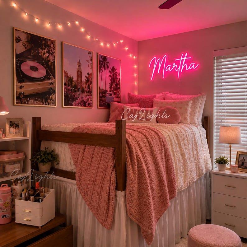 May include: A bedroom featuring a wooden loft bed with a white bed skirt, a pink textured throw blanket, and a pink neon sign displaying the name Martha. The room includes three framed wall prints, string lights, and a white bedside table lamp.
