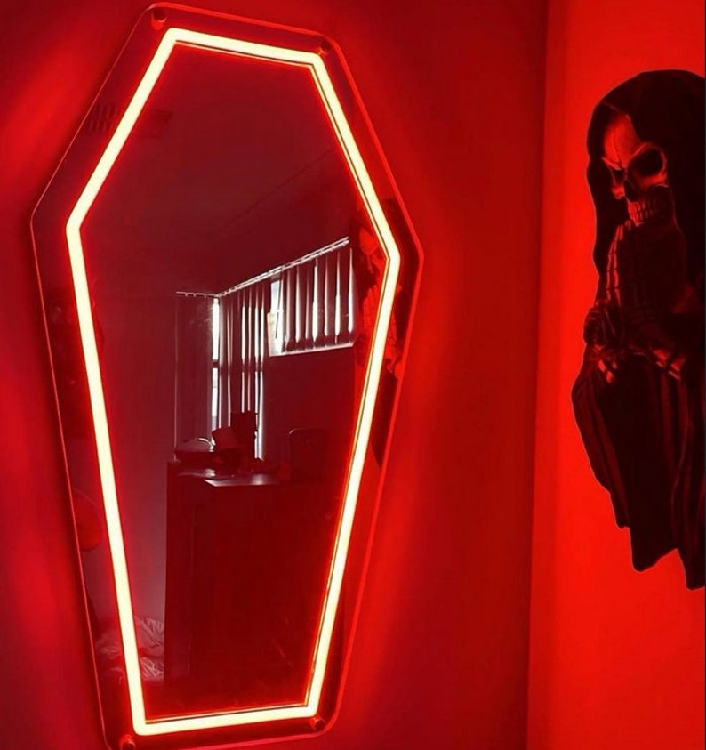 May include: A coffin-shaped mirror with a bright red neon border. The mirror is mounted on a red wall, and a decorative skull figure is to the right. The room is dimly lit, creating a spooky atmosphere.