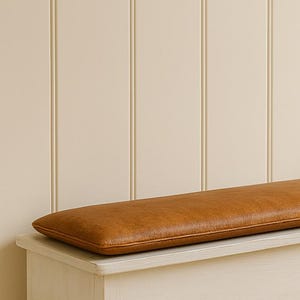 May include: A white wooden storage bench with a rectangular, brown leather cushion. The bench has four short, tapered legs and a simple, clean design. The cushion is a warm, medium brown color.