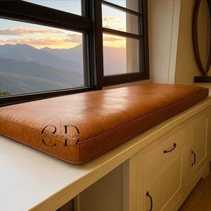 May include: A rectangular, brown leather window seat cushion with a stitched border. The cushion is placed on a white cabinet, with a window in the background overlooking a mountain range. The cushion is approximately 60 inches long.