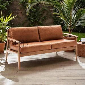 Op de afbeelding: Een tweezits loungesofa voor buiten met een lichtbruin houten frame en bruine leren kussens. De bank heeft een modern design met armleuningen en staat op een houten terras. Planten en een bakstenen muur zijn op de achtergrond.