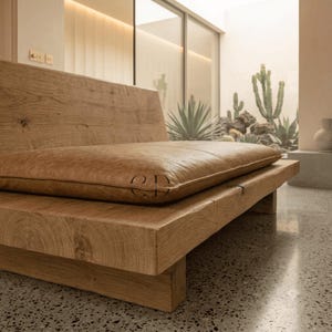 May include: A low-profile wooden bench featuring a thick, light brown leather cushion. The frame is constructed from solid, light-toned wood with visible grain patterns. The cushion is rectangular and smooth, featuring the letters CD embossed on it.