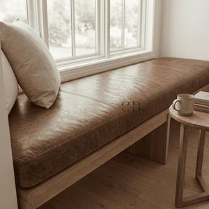 May include: A long bench featuring a brown leather cushion with a visible grain texture, supported by a light-toned wooden base. Two beige throw pillows rest on the left side, and a small wooden side table with a ceramic mug sits to the right.