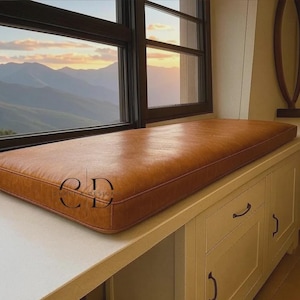 May include: A rectangular tan leather bench cushion with piped edges, positioned on a white built-in cabinet surface. The cushion features a smooth, matte texture and a consistent warm brown tone, resting directly beneath a large window frame.