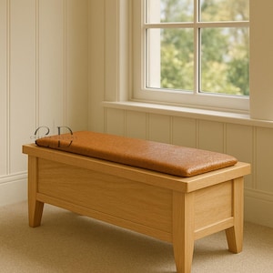 May include: A wooden storage bench with a light brown leather cushion. The bench has a rectangular shape with a sloped front and four legs. The bench is placed near a window.